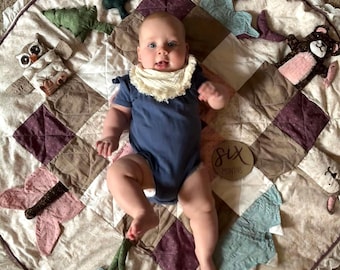 May include: A round, quilted play mat featuring a patchwork design in shades of cream, brown, and plum, accented with lace trim. The mat includes attached plush elements like an owl, trees, a whale, and a bear. A wooden disc reads six months.