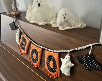 May include: A decorative Halloween banner featuring orange fabric pennants with black polka-dot letters spelling BOO, accented with small orange pom-poms. The banner includes hanging fabric ghost and black bat ornaments attached to a white lace trim.