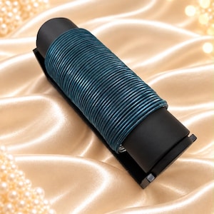 Op de afbeelding: Een set van talloze dunne, donker-türkis metalen armbanden gestapeld op een zwarte cilindrische displaystandaard. De armbanden hebben een glad, afgerond profiel en een consistente blauwgroene metallic afwerking, gerangschikt in een kolom.