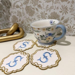 Puede incluir: Una taza de té de cerámica con estampado floral y asa azul claro, acompañada de tres posavasos de tela blanca con borde dorado festoneado y una letra S bordada en azul en el centro, dispuestos sobre una superficie plana.