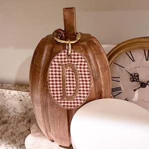 May include: A brown, ribbed, pumpkin-shaped decorative object featuring a gold-toned metal ring at the top. Attached to the ring is an oval-shaped fabric tag with a red and white gingham pattern and a large, embroidered letter D in the center.