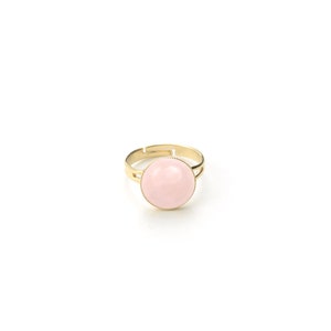 May include: A gold-toned adjustable ring featuring a large, round, light pink stone. The ring has a simple, elegant design with a smooth, polished finish. The stone is set in a delicate bezel setting, creating a classic and timeless look.
