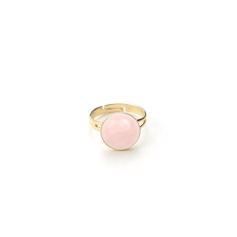 May include: A gold-toned adjustable ring featuring a large, round, light pink stone. The ring has a simple, elegant design with a smooth, polished finish. The stone is set in a delicate bezel setting, creating a classic and timeless look.