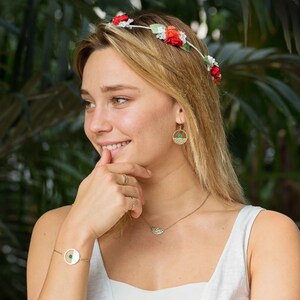 Pu&ograve; includere: Una donna indossa una canotta bianca con una collana e orecchini d'oro. Indossa una corona di fiori con fiori rossi e bianchi. Sorride e guarda di lato.
