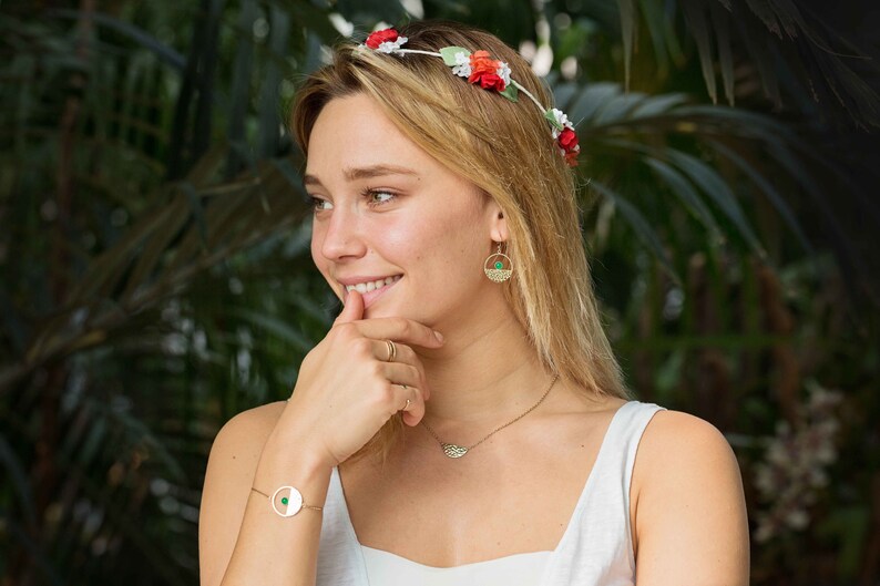 Pu&ograve; includere: Una donna indossa una canotta bianca con una collana e orecchini d'oro. Indossa una corona di fiori con fiori rossi e bianchi. Sorride e guarda di lato.