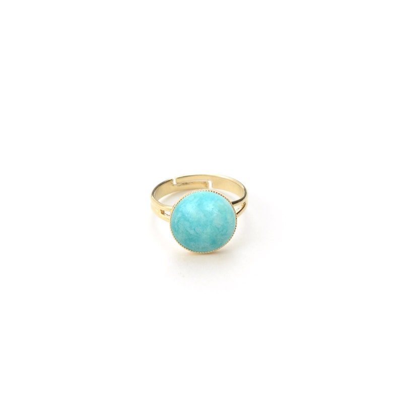 May include: A gold-toned adjustable ring featuring a large, round, light blue stone. The stone is set in a bezel setting with a textured edge. The ring is displayed on a white background.
