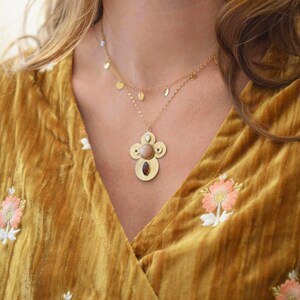 May include: A gold necklace with a large, ornate pendant featuring a sunstone and other gemstones. The pendant is surrounded by a circle of smaller stones. The necklace is worn by a person with long brown hair and a yellow velvet top.