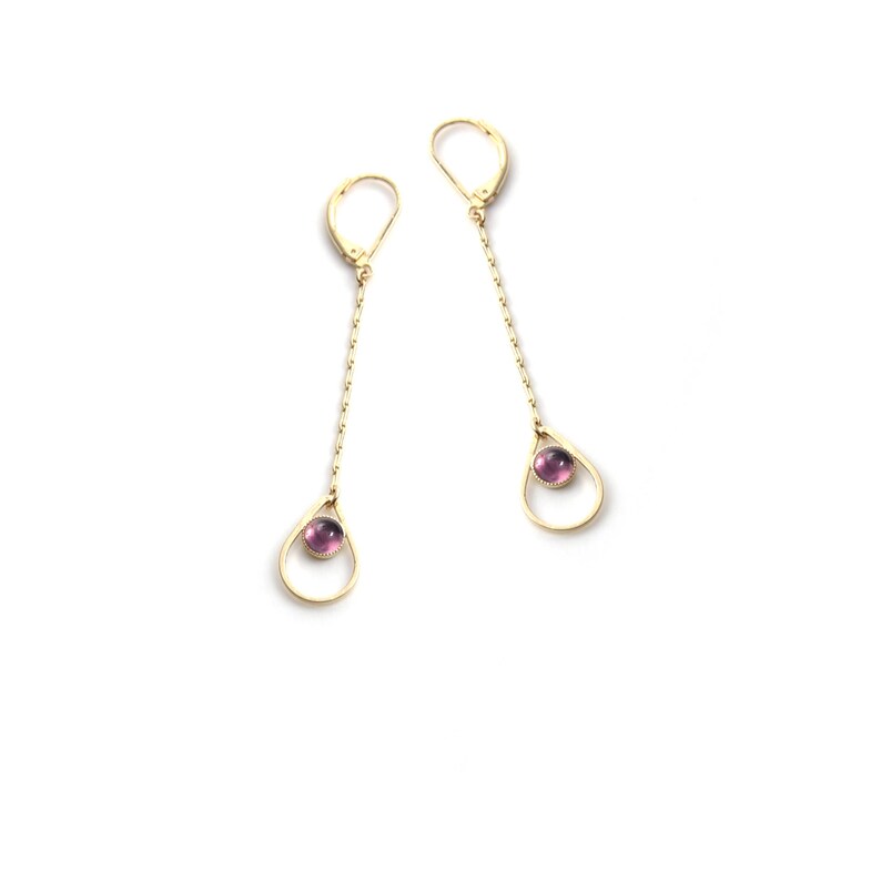 K&ouml;nnte beinhalten: Ein Paar goldene Ohrringe mit einem tropfenf&ouml;rmigen Anh&auml;nger und einem kleinen violetten Edelstein in der Mitte.