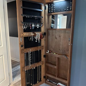 May include: A wall-mounted wooden jewellery armoire with a dark brown finish, featuring an open door with black velvet lining for hanging necklaces and earrings. The interior includes a mirror, LED lighting, ring slots, and multiple storage shelves.