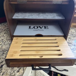 May include: A dark brown wooden bread box with a hinged lid and a pull-out bamboo cutting board base. The interior features two shelves, one with a patterned design and the other with the text LOVE IN EVERY SLICE printed in black on a white surface.