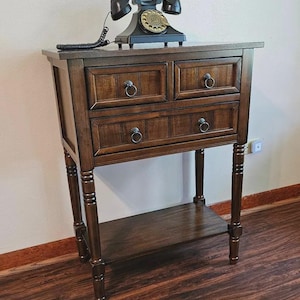 May include: A dark brown wooden console table with two small top drawers, one wide bottom drawer, and a lower shelf. The table features four turned legs and round metal ring pulls. A black vintage rotary telephone sits on the tabletop.
