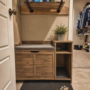 May include: A wooden storage bench with a gray seat cushion, a side shelving unit, and a black metal wall shelf above. A gray mat with tan paw prints lies on the floor. The bench features a cabinet door and an open shelf for storage.