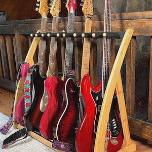 May include: A light-colored wooden guitar stand holding five electric guitars and basses. The instruments feature various finishes including solid red, sunburst, and wood grain, with black pickguards and patterned fabric straps attached to the bodies.