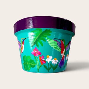 Puede incluir: Una maceta de color turquesa con un borde morado oscuro, pintada a mano con dos colibríes en vuelo, hojas tropicales de color verde vibrante y varias flores coloridas, incluyendo flores rosas y margaritas blancas sobre un fondo azul.