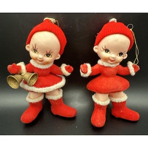 May include: Two vintage-style doll ornaments wearing red felt outfits with white faux fur trim and red knit hats. The dolls have pale skin, large eyes, and red boots. One doll holds two gold-coloured bells, while the other has a gold hanging loop.