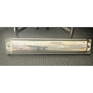 May include: A silver-toned metal harmonica featuring the engraved text THE GOLIATH and MADE BY M. HOHNER GERMANY on the front cover plate. The instrument has a rectangular shape with rounded ends and visible screws on the left and right sides.
