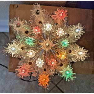Op de afbeelding: Een stervormige kerstboompiek met zilveren klatergoudfranjes en meerdere fittingen. De piek is verlicht met kleine lampjes in diverse kleuren, waaronder rood, oranje, groen, blauw en wit, wat zorgt voor een veelkleurige gloed.
