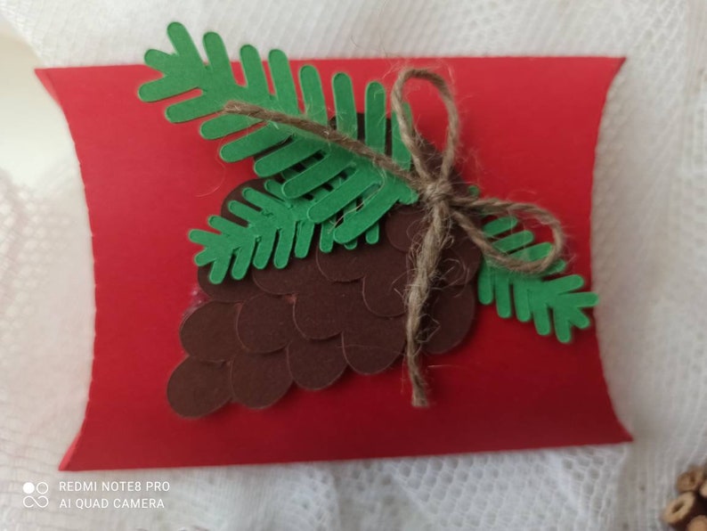 Peut inclure: Une bo&icirc;te &agrave; coussin rouge d&eacute;cor&eacute;e d'une pomme de pin en papier marron et verte et d'une branche de pin. La pomme de pin est faite de cercles de papier superpos&eacute;s et la branche de pin est faite de papier vert.