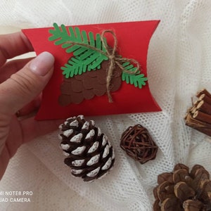 Peut inclure: Une bo&icirc;te coussin en feutre rouge avec un motif de pin en papier vert et de c&ocirc;ne de pin brun. La bo&icirc;te est pos&eacute;e sur une surface en dentelle blanche.