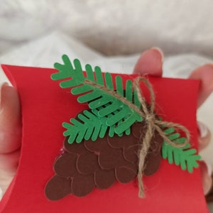 Peut inclure: Une bo&icirc;te &agrave; coussin rouge avec un c&ocirc;ne de pin brun et des aiguilles de pin vertes sur le dessus. La bo&icirc;te est attach&eacute;e avec de la ficelle.