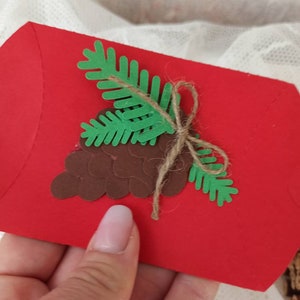 Peut inclure: Une enveloppe rouge d&eacute;cor&eacute;e d'une pomme de pin brune et d'une branche de pin verte. L'enveloppe est attach&eacute;e avec de la ficelle.