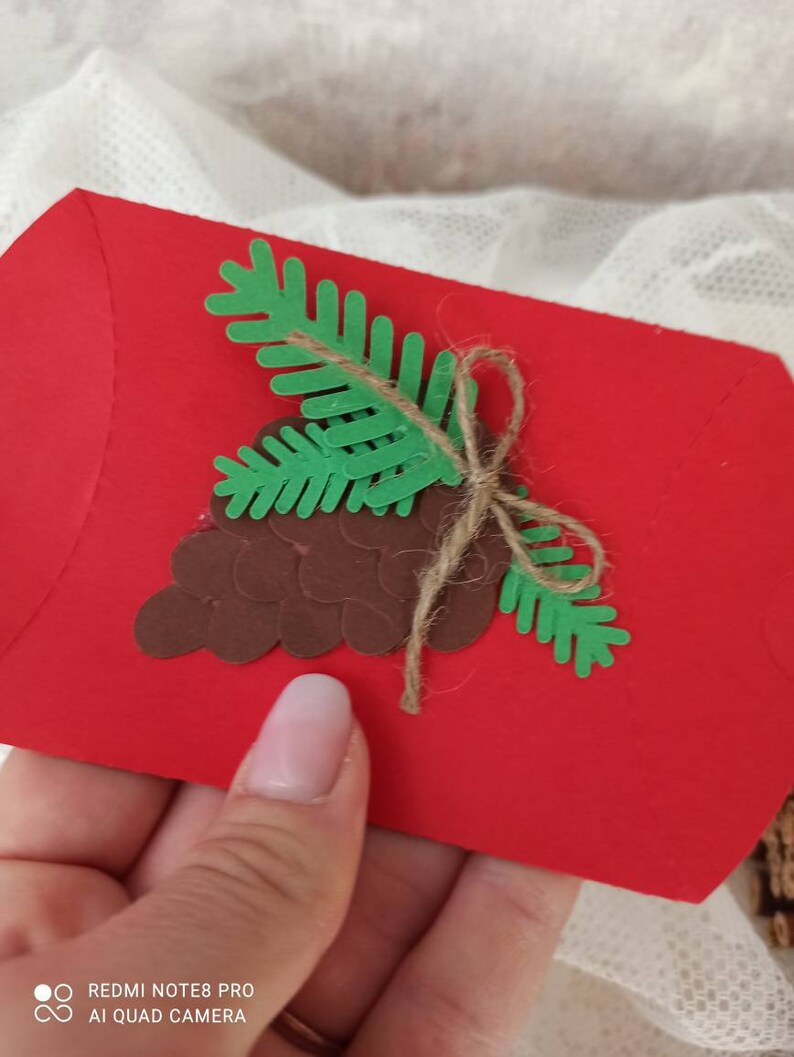 Peut inclure: Une enveloppe rouge d&eacute;cor&eacute;e d'une pomme de pin brune et d'une branche de pin verte. L'enveloppe est attach&eacute;e avec de la ficelle.