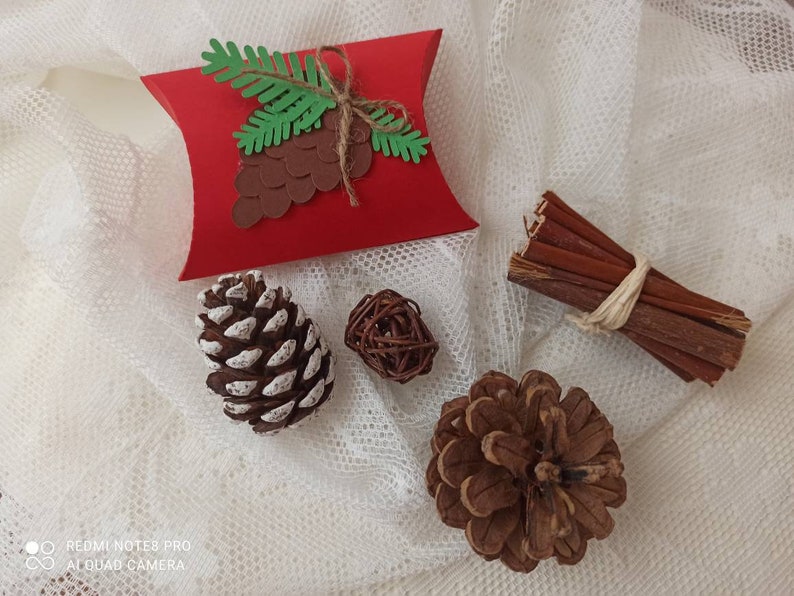 Peut inclure: Une bo&icirc;te &agrave; coussin rouge avec un motif de branche de pin verte et de c&ocirc;ne de pin brun. La bo&icirc;te est sur un fond de tissu en dentelle blanche avec un c&ocirc;ne de pin brun, un petit fagot de brindilles brunes et une boule en osier brune.
