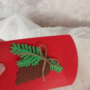 Peut inclure: Un sac cadeau en papier rouge d&eacute;cor&eacute; d'une pomme de pin brune et d'une branche de pin verte attach&eacute;e avec de la ficelle.
