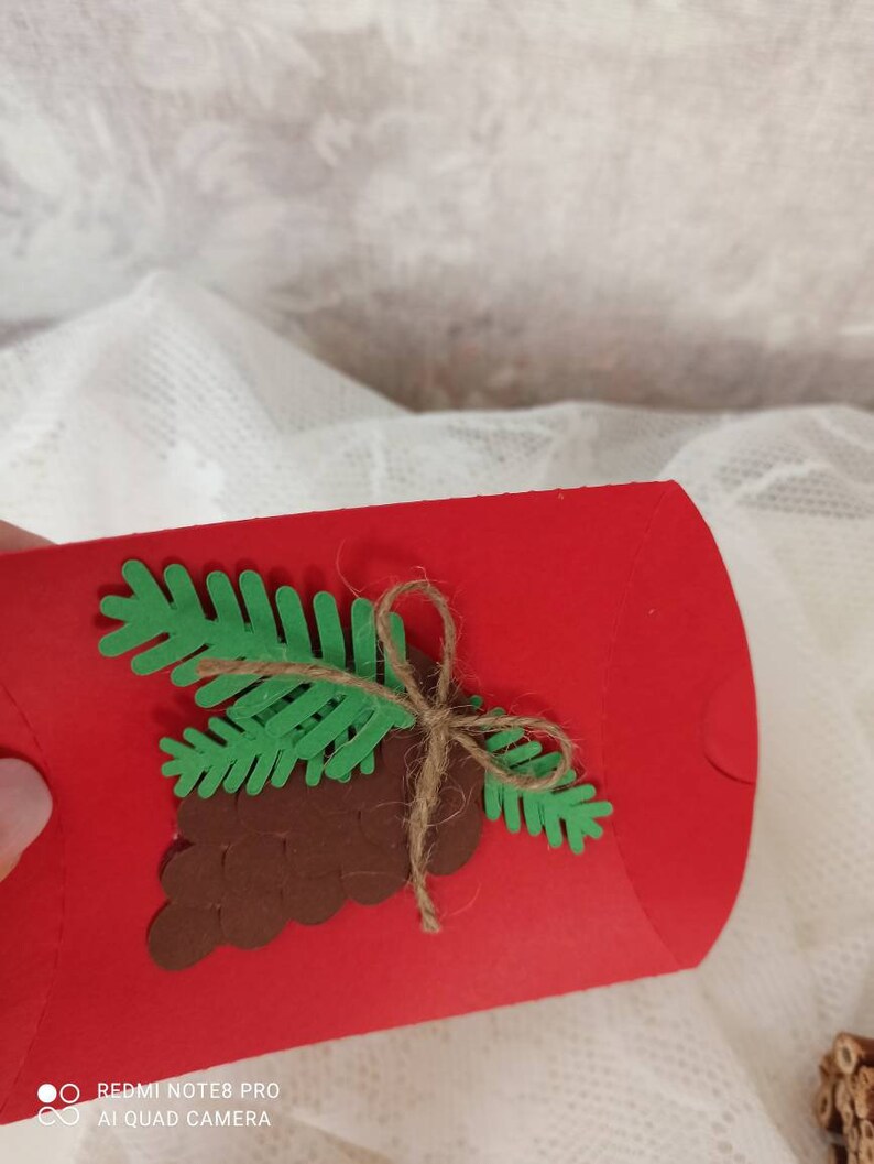 Peut inclure: Un sac cadeau en papier rouge d&eacute;cor&eacute; d'une pomme de pin brune et d'une branche de pin verte attach&eacute;e avec de la ficelle.