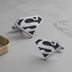 May include: A pair of silver-toned metal cufflinks, each shaped like a diamond-style shield with a black enamel inlay forming the letter S. The design features a polished metallic border and a classic toggle backing for secure attachment to shirt cuffs.