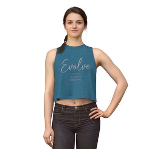 May include: A teal sleeveless crop top featuring the word Evolve in a cursive script font, with the words GROW, BLOOM, and BECOME printed in a smaller, sans-serif font directly underneath. The garment is paired with dark charcoal denim jeans.