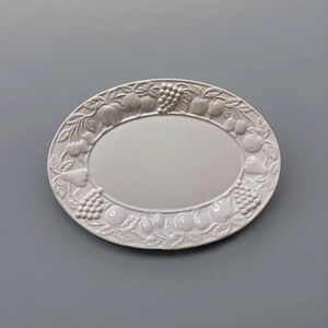 May include: An oval-shaped white ceramic platter featuring a raised, embossed border design of various fruits including grapes, pears, cherries, and apples. The center of the platter is smooth and plain, while the rim is decorated with detailed fruit motifs.