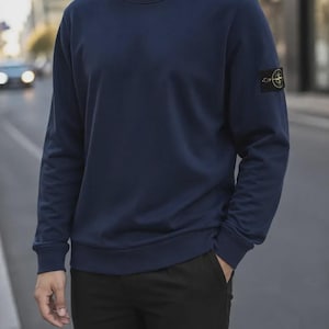 May include: A navy blue long-sleeve crewneck sweatshirt with a ribbed collar, cuffs, and hem. The left sleeve features a black rectangular patch with a yellow and green compass-style logo emblem embroidered on the centre of the patch.