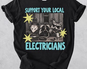 Puede incluir: Una camiseta negra con un gráfico de tres animales trabajando con herramientas eléctricas. El texto dice SUPPORT YOUR LOCAL ELECTRICIANS en azul claro. La imagen incluye una foto en blanco y negro de una zarigüeya, una mofeta y un mapache con gafas de sol, acentuada por formas de estrellas amarillas.