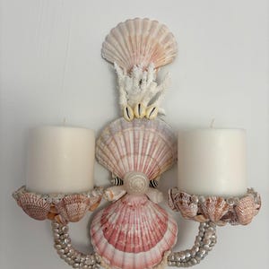 May include: A wall-mounted candle holder crafted from various seashells and white coral. The central structure features stacked pink and white scallop shells, while two curved arms support circular platforms covered in small brown shells, each holding a white pillar candle.