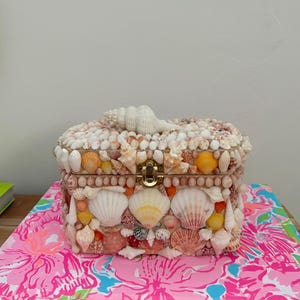 Könnte beinhalten: Eine rechteckige Schatulle, die mit verschiedenen Muscheln in Weiß-, Creme-, Rosa- und Orangetönen bedeckt ist. Der Deckel hat eine große, längliche weiße Spiralmuschel als Griff. Ein goldfarbener Metallverschluss sichert die Vorderseite der mit fächerförmigen und spiralförmigen Muscheln verzierten Schatulle.