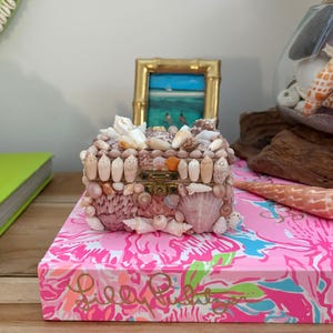 May include: A rectangular keepsake box covered in various small white, tan, and pink seashells. The box features a brass-colored metal latch on the front and is decorated with a dense arrangement of shells of different shapes and sizes.