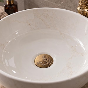 Peut inclure: Une vasque ronde en céramique blanche avec un motif subtil rappelant le marbre beige. Le lavabo est doté d'une bonde circulaire en laiton gravée d'un motif de branche florale, accompagnée d'un robinet en laiton antique ornementé.