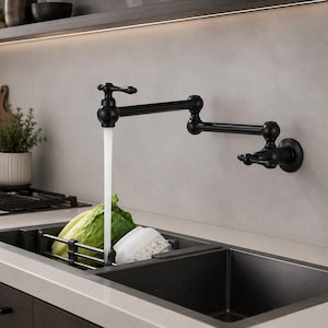 Può includere: Un rubinetto a parete nero opaco per riempire pentole, dotato di braccio snodabile doppio e due manopole, esteso sopra un lavello da cucina scuro. L'acqua scorre dall'erogatore in uno scolapiatti in metallo nero contenente lattuga e una ciotola bianca.