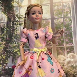 May include: A doll wearing a pink dress with a yellow ribbon and a white tulle underskirt. The dress features a pattern of cartoon princesses. The doll has blonde hair and is wearing pink shoes.