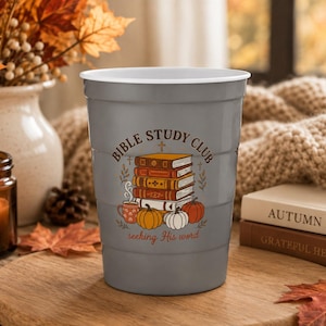 May include: A gray plastic stadium cup featuring a graphic of a stack of books, pumpkins, and a mug. The text reads BIBLE STUDY CLUB and seeking His word. The design includes small crosses and foliage accents printed in brown, orange, and white.