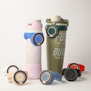 May include: Two water bottles, one light pink and one olive green, featuring integrated carry loops and flip-top lids. The green bottle displays the text FUEL THE BODY BUILD THE LIFE. Several additional silicone carry loops in various colors are arranged.