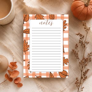 Op de afbeelding: Een rechthoekig notitieblok met een wit middenstuk met horizontale zwarte lijnen en het woord notes in cursief schrift bovenaan. De rand heeft een oranje en wit ruitpatroon, versierd met diverse illustraties van bruine herfstbladeren.