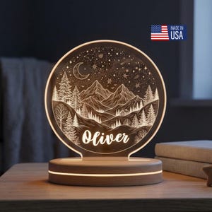 May include: A circular LED night light featuring an engraved mountain landscape with pine trees, a crescent moon, and a starry sky. The name Oliver is etched in white script at the bottom center. The light sits on a round, light-colored wooden base.