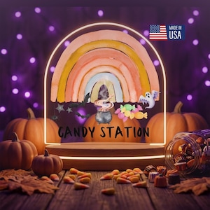 May include: An arched acrylic sign featuring a watercolor rainbow, a cauldron with smoke, bats, a ghost, and assorted candies. The text reads CANDY STATION. The sign is mounted on a wooden base and includes a small Made in USA flag icon.