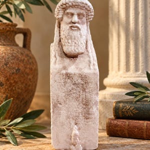 Puede incluir: Una escultura de hermes color piedra que presenta una cabeza masculina barbuda con un gorro texturizado y cabello largo, sobre una base de pilar rectangular. La superficie tiene un acabado mate desgastado con sutiles reflejos marrones.