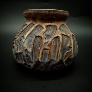 Könnte beinhalten: Eine kleine, runde Keramikvase mit einem breiten, ausgestellten Rand. Die Außenseite weist ein tief strukturiertes, geometrisches Reliefmuster mit einem gesprenkelten Finish in erdigen Braun-, Beige- und gedämpften Blautönen auf, was eine raue Oberfläche erzeugt.