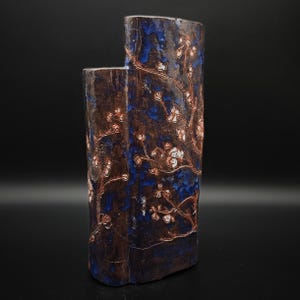 May include: A tall, cylindrical ceramic vase with an uneven top edge, featuring a dark brown and deep blue mottled glaze. The surface is decorated with a raised, hand-painted pattern of delicate white and light pink flowering tree branches.