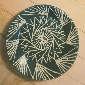 May include: A round ceramic plate featuring a deep forest green glaze with a contrasting white hand-painted geometric pattern. The design consists of a central sunburst motif surrounded by repeating triangular shapes and radiating lines.