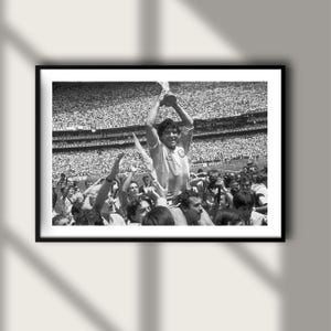 May include: A black and white photograph depicting a football player being carried on shoulders in a crowded stadium, holding a trophy high above their head. The scene captures a celebratory moment with numerous spectators and photographers in the background.